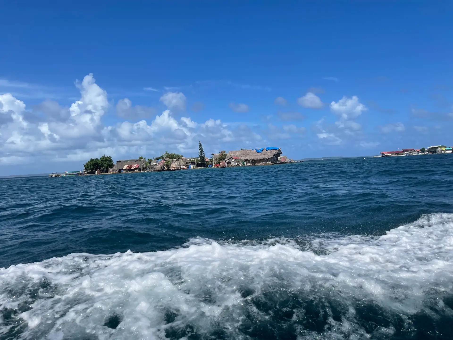 Photo of one of the residential San Blas islands, taken from the boat ferry. It is a crowded collection of shanties with thatched roofs, some of which have blue tarps over parts of them.