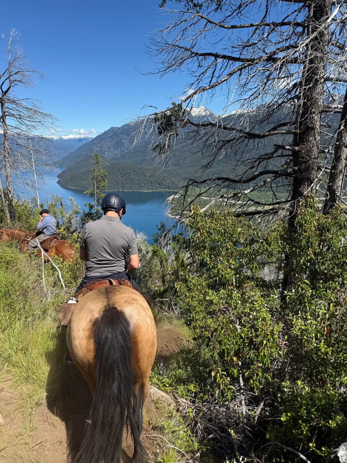 The backs of two riders on horses making their way around a mountain ride overlooking Lago Puelo